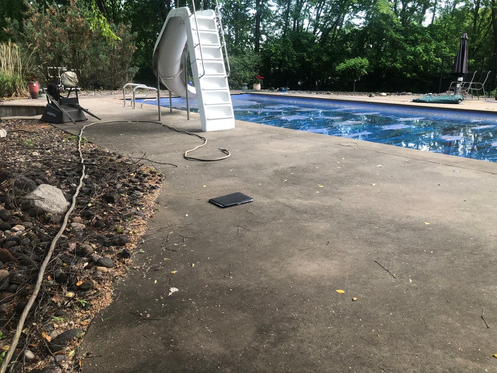 Swimming pool area with slide, chair, and electric cable on concrete surface among greenery.
